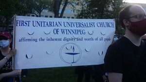 photo of man in mask at a protest, standing in front of First Unitarian Universalist Church of Winnipeg banner